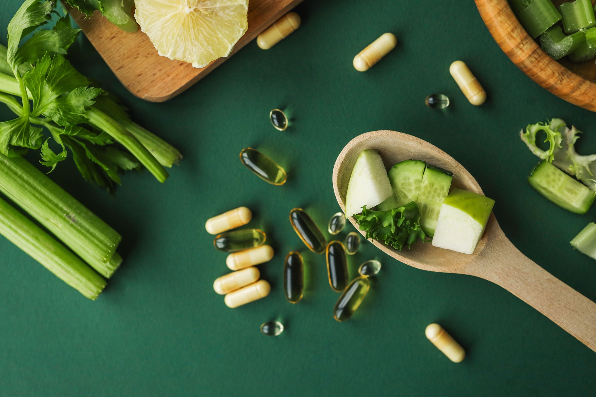 Overhead view of chopped celery, cucumber, and lemon alongside vitamin capsules on a wooden board against a green background. Perfect for healthy lifestyle or detox meal prep